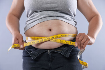 Overweight woman measuring her belly with tape on light background, closeup