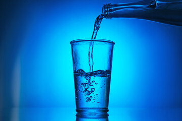 Pouring water from bottle into glass on blue gradient background. Backlit
