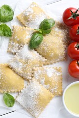 Uncooked ravioli, basil, tomatoes and oil on white wooden table, flat lay