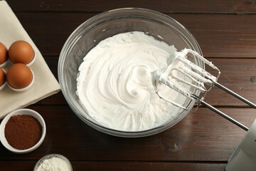 Bowl with whipped cream, hand mixer and ingredients on wooden table, flat lay