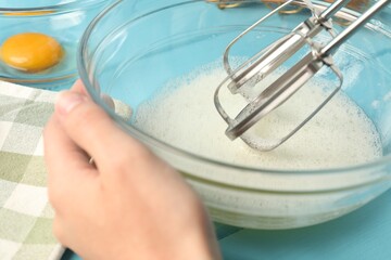 Woman whisking egg whites with hand mixer at light blue wooden table, closeup
