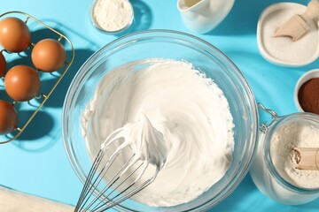 Whisk with whipped cream, eggs and other products on light blue table, flat lay