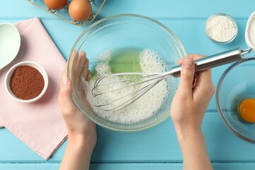 Woman whisking egg whites in bowl at light blue wooden table, top view