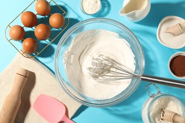 Whisk with whipped cream, eggs and other products on light blue table, flat lay