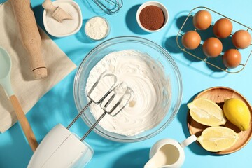Hand mixer with whipped cream, eggs and other products on light blue table, flat lay