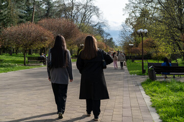 Two people stroll through a public park lined with trees and benches on a sunny day in springtime
