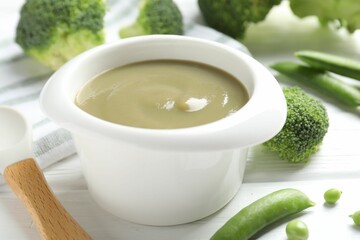 Baby food. Tasty puree in bowl, spoon, peas and broccoli on white wooden table, closeup