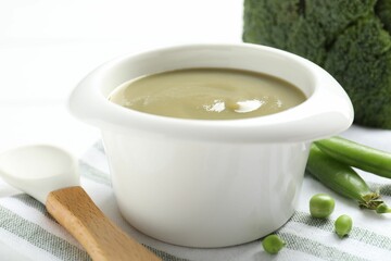 Baby food. Tasty puree in bowl, spoon, peas and broccoli on white table, closeup