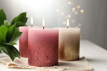Burning candles and green leaves on white table against blurred lights, closeup