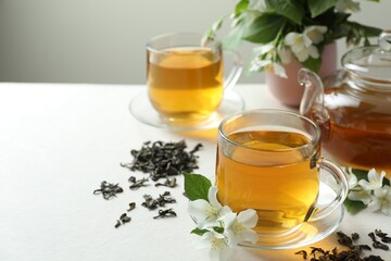 Aromatic jasmine tea in glass cups, teapot, flowers and dried leaves on white table, closeup. Space for text