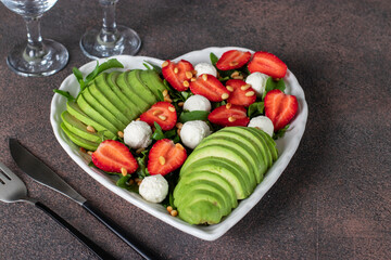 Healthy salad with strawberries, avocado, arugula and feta cheese in heart-shaped plate on brown...