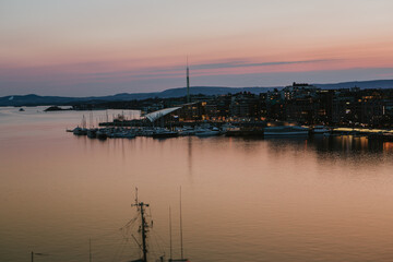 A vibrant sunset bathes the Astrup Fearnley Museum of Modern Art in Oslo, with a ferry gracefully sailing by, creating a harmonious blend of art, architecture, and the beauty of the Oslofjord