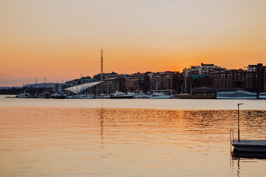 Bathed in the warm glow of sunset, Oslo harbour showcases traditional colourful wooden houses along the waterfront, with boats gently berthed in the distance.