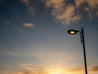A street LED light is lit up in the evening sky. The sky is mostly clear with a few clouds scattered around. The light is shining brightly, illuminating the area below. The scene has a peaceful mood