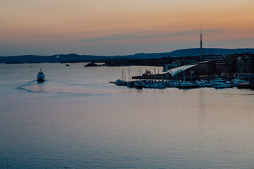 A vibrant sunset bathes the Astrup Fearnley Museum of Modern Art in Oslo, with a ferry gracefully sailing by, creating a harmonious blend of art, architecture, and the beauty of the Oslofjord