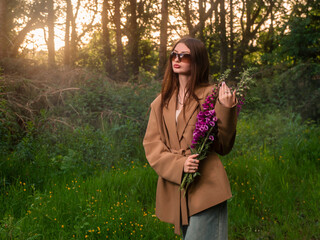 Young stylish girl in brown coat and sun glasses holding a bouquet of wild purple flowers. The...