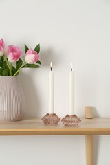 Elegant holders with burning candles, tulips in vase and other decor on wooden table near white wall