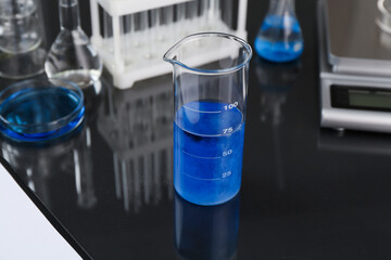 Beaker with blue liquid, other laboratory glassware and scale on black mirror table, closeup