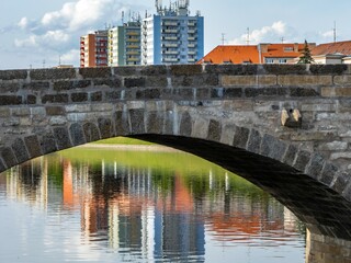 Naklejka premium Colorful apartment buildings reflected in calm water beneath a stone bridge arch. Urban landscape with contrast between historical architecture and modern housing