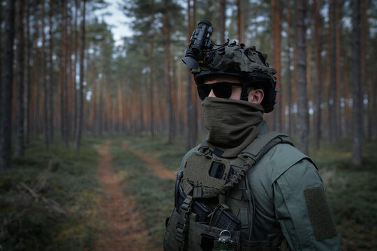An elite unit soldier in full tactical gear and a helmet with a night vision device conducts surveillance during a patrol in the forest. - Powered by Adobe