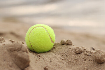 Yellow tennis ball lies on the sand. Sports and recreation concept, summer games. Tennis or paddleball ball. Tennis court near the beach. Paddleball ball on the sand near the water. Outdoor games