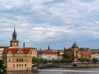 Historic buildings and towers along the Vltava River in Prague under a cloudy sky. Classic European architecture with red rooftops and waterfront charm