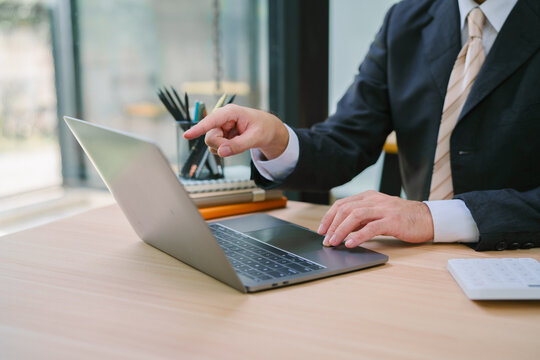 Asian businessman focused and determined, sitting at a marble-top desk with a sleek laptop in a modern workspace, a potted plant, and a floating shelf with books