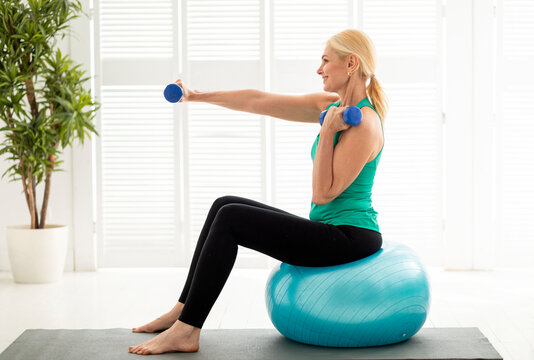 Sporty mature woman working out with dumbbells on fitness ball at home, fit senior female exercising in living room interior, enjoying domestic trainings and active lifesyle, side view, copy space