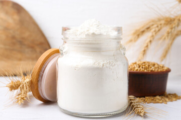 Wheat flour in glass jar, grains and spikes on white table, closeup