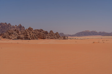Desert Landscape under Blue Sky: An expansive view of a desolate desert, featuring vast stretches of orange sand meeting the rugged, imposing rock formations under a cloudless, azure sky.