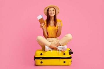 Emotional young woman in summer hat holding passport and plane tickets, sitting on big yellow luggage, ready to travel. Cheerful lady traveller posing over pink studio background, ready for vacation © Prostock-studio