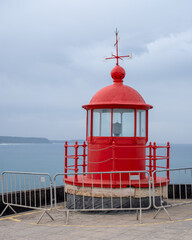 Bright red lighthouse structure above stone platform with ocean and cloudy sky in background.