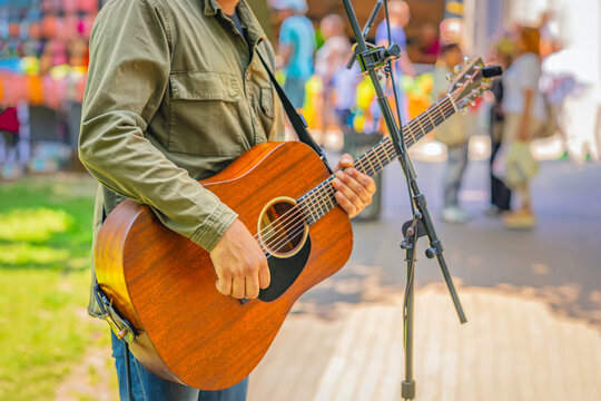 Man playing acoustic guitar on stage during outdoor event, surrounded by people and colorful background. Concept of live music, busking, entertainment and street performance energy