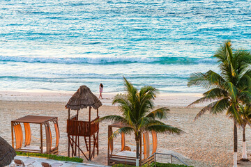 Woman walking along a serene beach in Cancun, surrounded by palm trees, wooden cabanas, and gentle ocean waves High-quality photo
