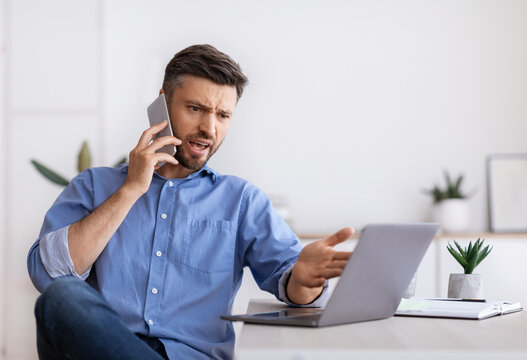 Angry millennial businessman talking on mobile phone and looking at laptop screen working in modern office, having problems with project, annoyed boss speaking with employee by phone, copy space