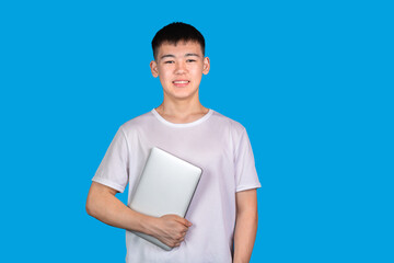 Smiling asian young man teenager in white T-shirt holding modern laptop against blue background