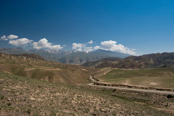 Wide angle landscape of a winding road going through a highland mountain pass surrounded by high peaks with glaciers. Summer day, blue sky with white clouds. Kyrgyzstan, Naryn Region, Central Asia.