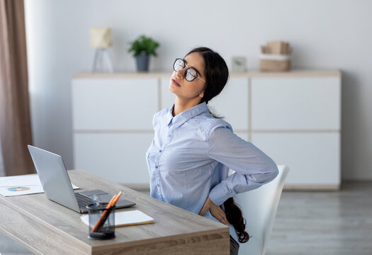 Fototapeta Tired upset young indian business woman suffering from back pain, muscle spasm at workplace with laptop in office or home interior. Fatigue, deadline, pain and incorrect posture, work at computer