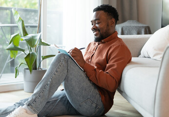 Cheerful Black Male Writer Writing And Taking Notes In Notebook Sitting On Floor At Home On...