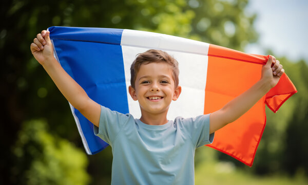 Joyful boy holding large French flag overhead outdoors. Child celebrating Bastille Day in a park. French language concept