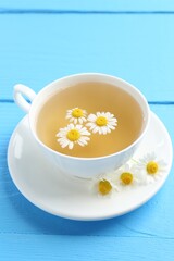 Delicious chamomile tea in cup and fresh flowers on light blue wooden table, closeup