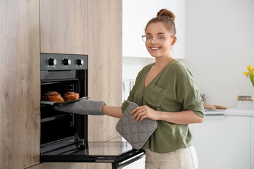 Happy young woman taking tray with fresh cinnamon rolls from electric oven