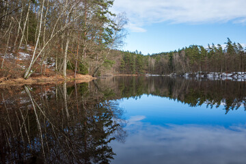 Barsjön lake with reflections of a mixed forest and a snowy bank under a blue sky in Vedema, Hässleholm, Sweden