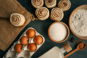 A top-down view of cinnamon buns on a dark surface with flour, sugar, eggs, milk, and a rolling pin, perfect for baking and recipe themes