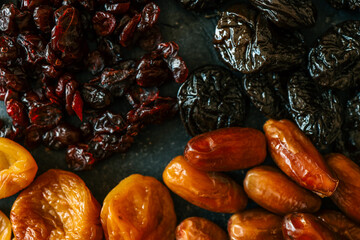 Four types of dried fruits-cranberries, dates, prunes, and apricots-arranged in rows on a dark textured background. A colorful and healthy assortment of natural snacks
