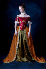 A red-haired Renaissance woman wearing an embroidered silk ensemble against a studio backdrop