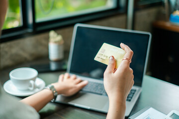 A woman uses a laptop and credit card for online shopping in a cozy cafe, with daylight streaming through the window.