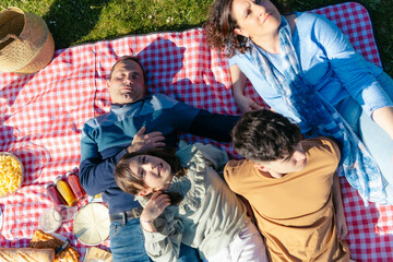 Fototapeta premium Happy family relaxing on a picnic blanket, enjoying food and sunshine in a park, spending quality time together