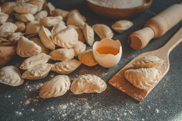 Pile of raw handmade dumplings with flour, cracked egg, rolling pin, and wooden board on dark rustic kitchen surface