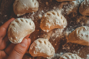 Homemade dumplings covered in flour on wooden table, close-up of hand holding raw pierogi before cooking
